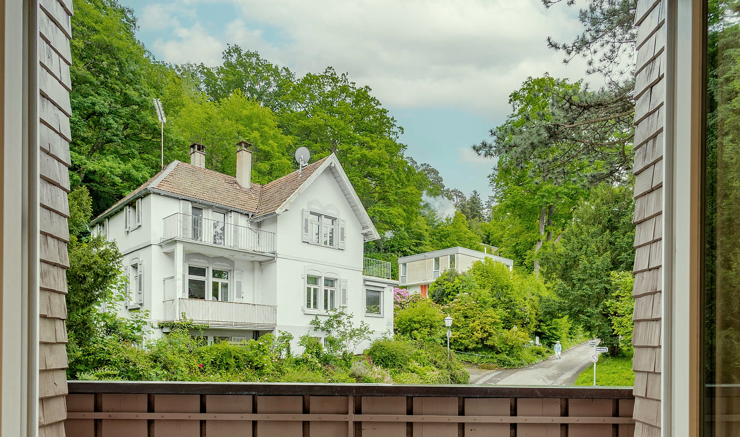 Blick von der Loggia eines Apartments: eine weiße Villa am bewaldeten Hang, davor Bäume und Sträucher